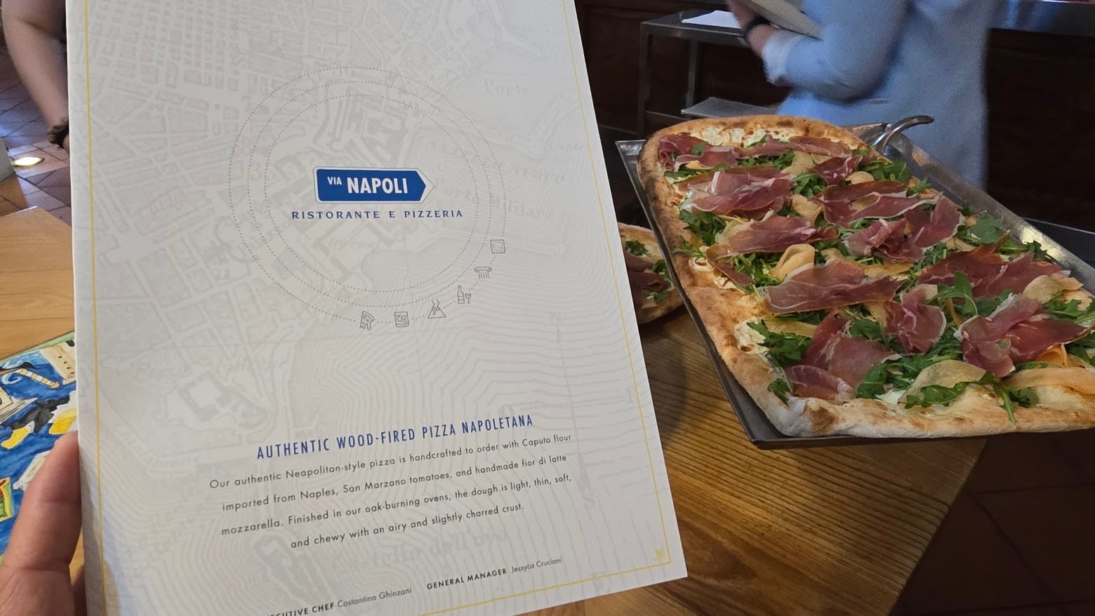 Person holds a Via Napoli menu next to a long rectangular pizza topped with prosciutto and arugula on a wooden table