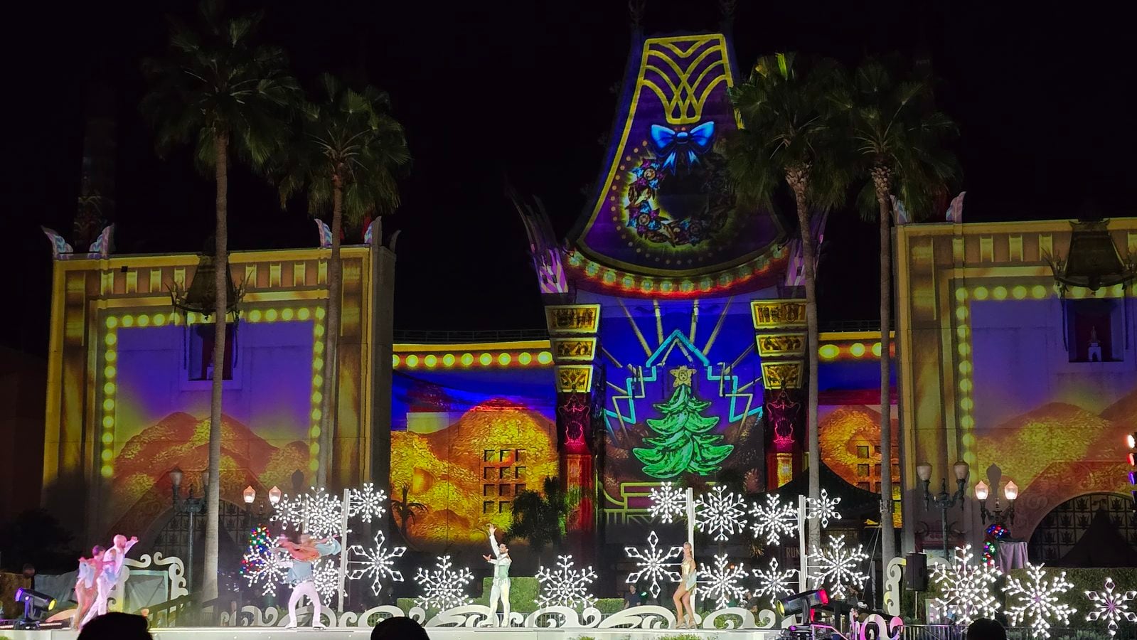 Nighttime carnival stage with colorful projection on a large arch shaped backdrop palm trees flanking the lit building and festive decorations on display