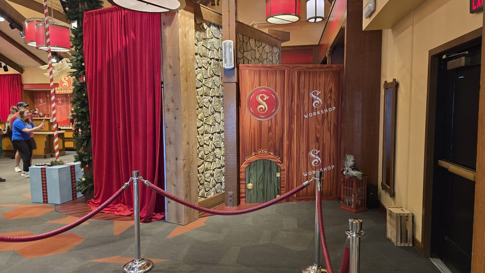 Indoor workshop set with red velvet curtains wooden posts stone wall and a decorative Workshop sign holiday decorations and people nearby in the background