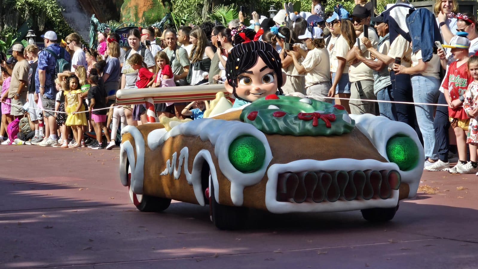 Crowd lines a parade route as a large colorful float shaped like a car with a cartoon character head passes by