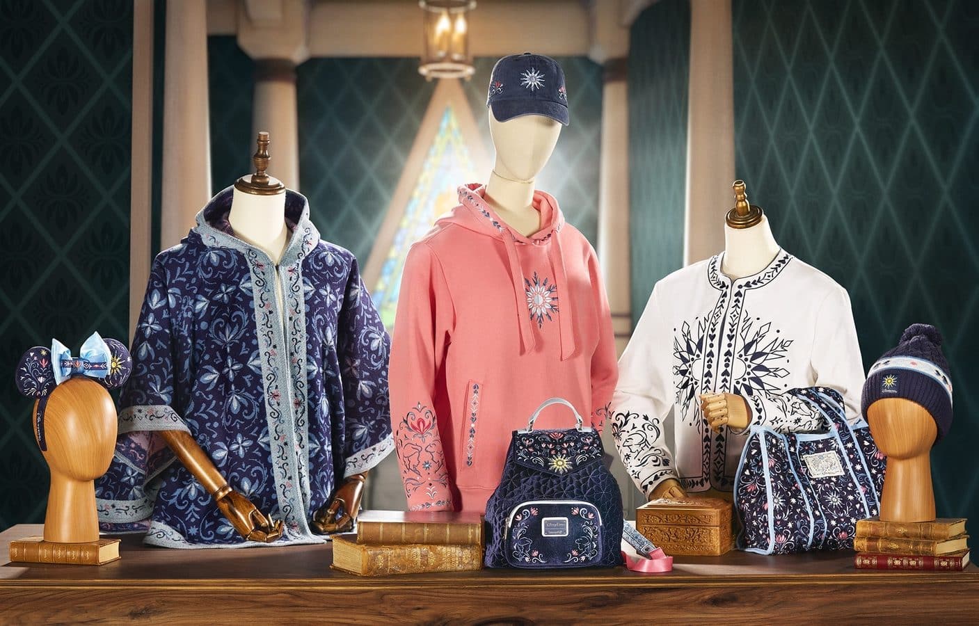 Shop window display with three mannequins wearing blue patterned robes and a pink embroidered hoodie plus hats bags and accessories on a wooden table
