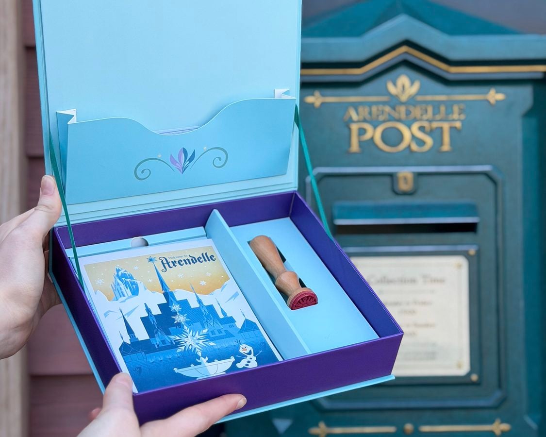 Person holds an open blue Advent calendar with a snowy village illustration near a blue post mailbox suggesting holiday mail delivery