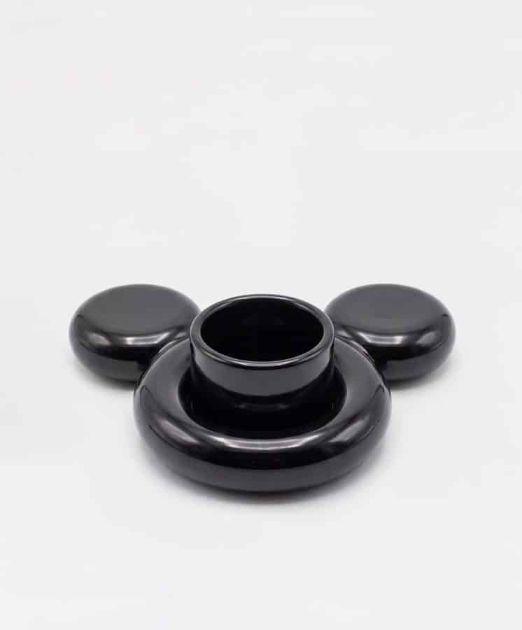 Black abstract ceramic sculpture with a central hollow cylinder and two rounded side knobs resting on a light surface