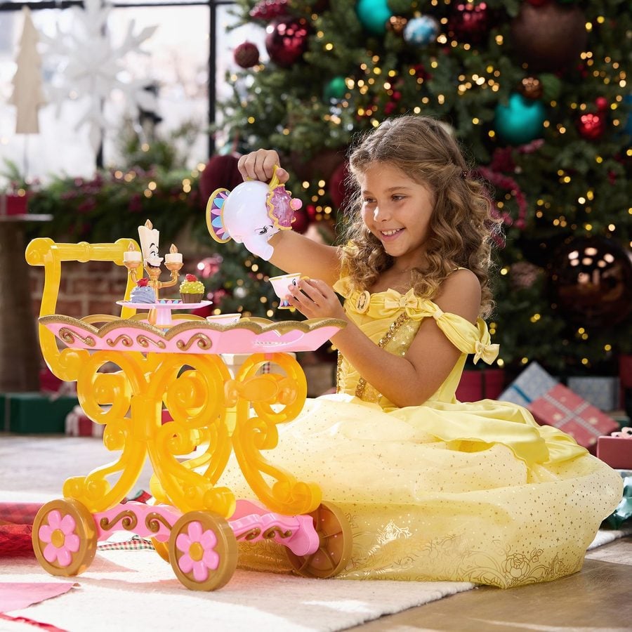 A girl in a yellow dress playing with a pastel themed toy cake stand and tea set beside a Christmas tree backdrop