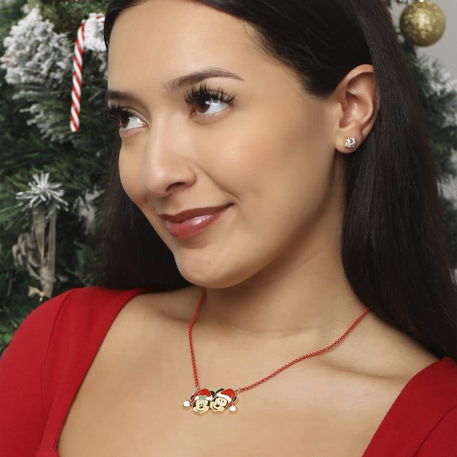 Close up of a woman in a red dress wearing a gold necklace with Mickey and Minnie Mouse charms set against Christmas tree decorations in the background