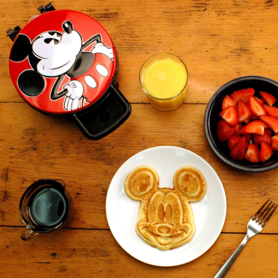Overhead view of a Mickey Mouse shaped waffle on a white plate with a glass of orange juice and a bowl of sliced strawberries on a wooden table