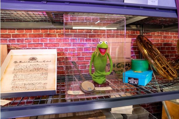 Kermit the Frog plush inside a clear display case on a shelf with a brick pattern backdrop flanked by a framed document and a brass horn display on the right