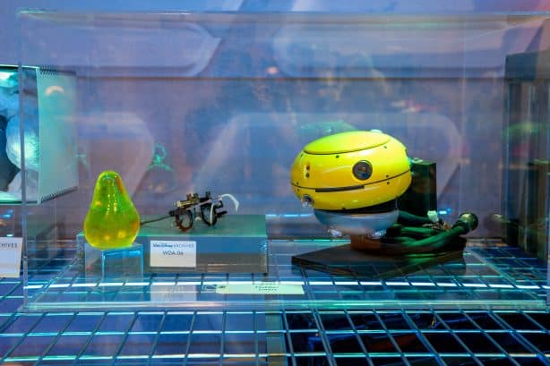 Display case with a yellow spherical robot a small model bicycle and a translucent green teardrop object under blue lighting part of a science exhibit