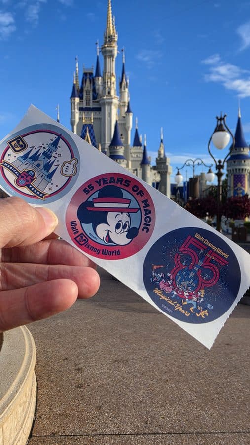 Person holding a sheet of Disney stickers in front of Cinderella Castle at Walt Disney World colorful icons celebrating 55 years of magic and park merch visible