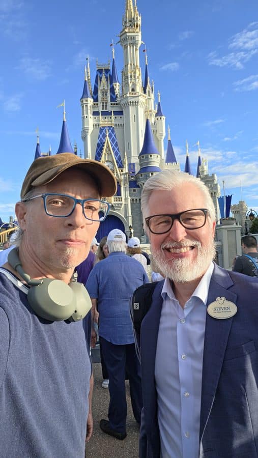 Selfie of two casually dressed men posing in front of a large blue and gray castle against a clear blue sky at a Disney park Left man wears cap and headphones right man in suit with name badge