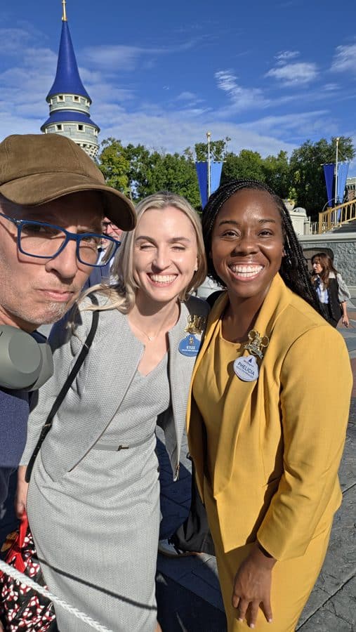 Group of three friends taking a sunny selfie outdoors near a castle like tower two women wear name badges and smile at the camera