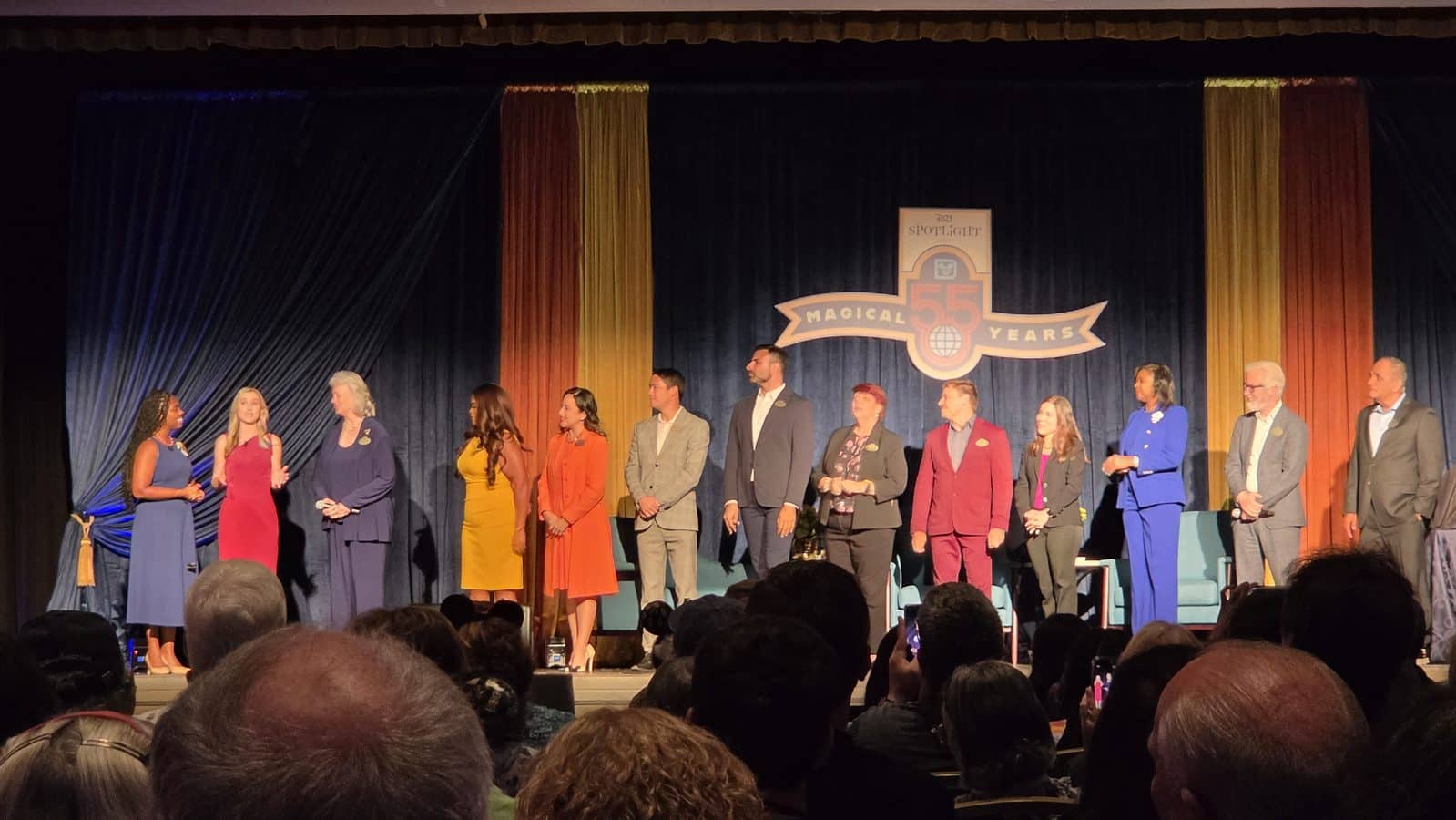 Group of people on a stage in colorful outfits facing the audience during a 55th anniversary celebration