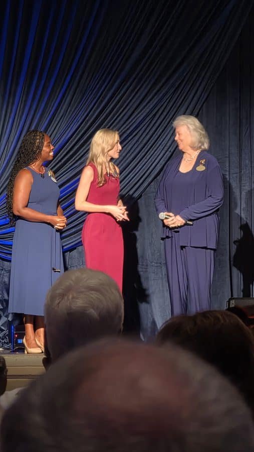 Three women in formal dresses on a stage an older woman in navy receives a microphone from two younger women in blue and magenta blue curtain backdrop