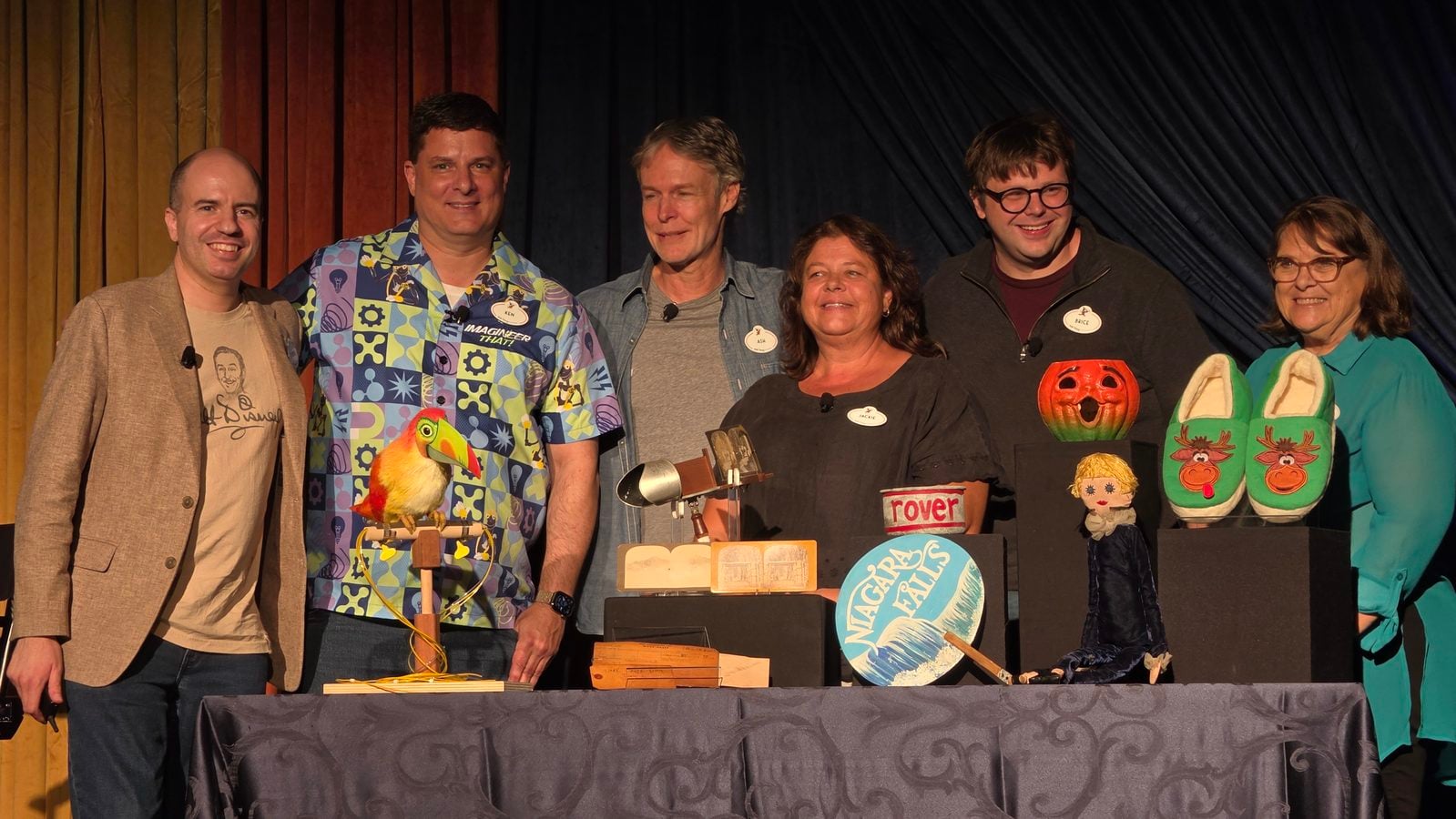 Group of six adults on a stage posing for a photo with crafts and props in front of a dark curtain backdrop