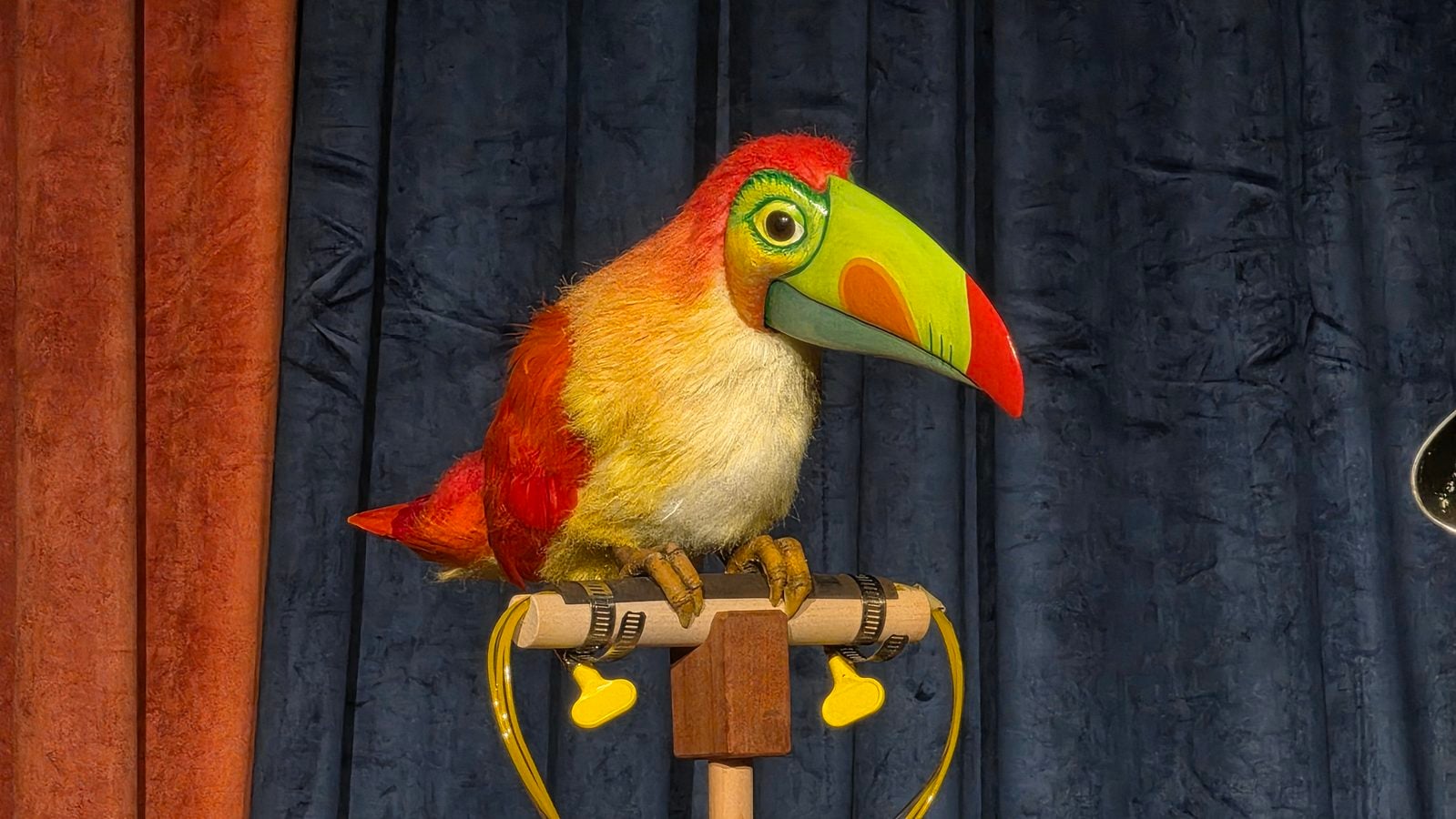 Colorful toucan puppet perched on a wooden stand in front of red and blue stage curtains for a performance