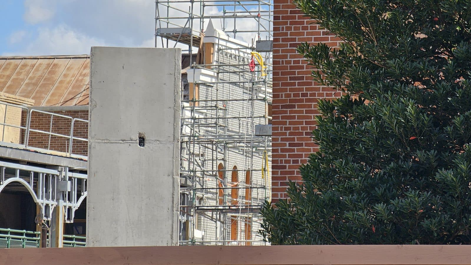Construction site with metal scaffolding beside a brick building and a tree on the right under a blue sky with clouds