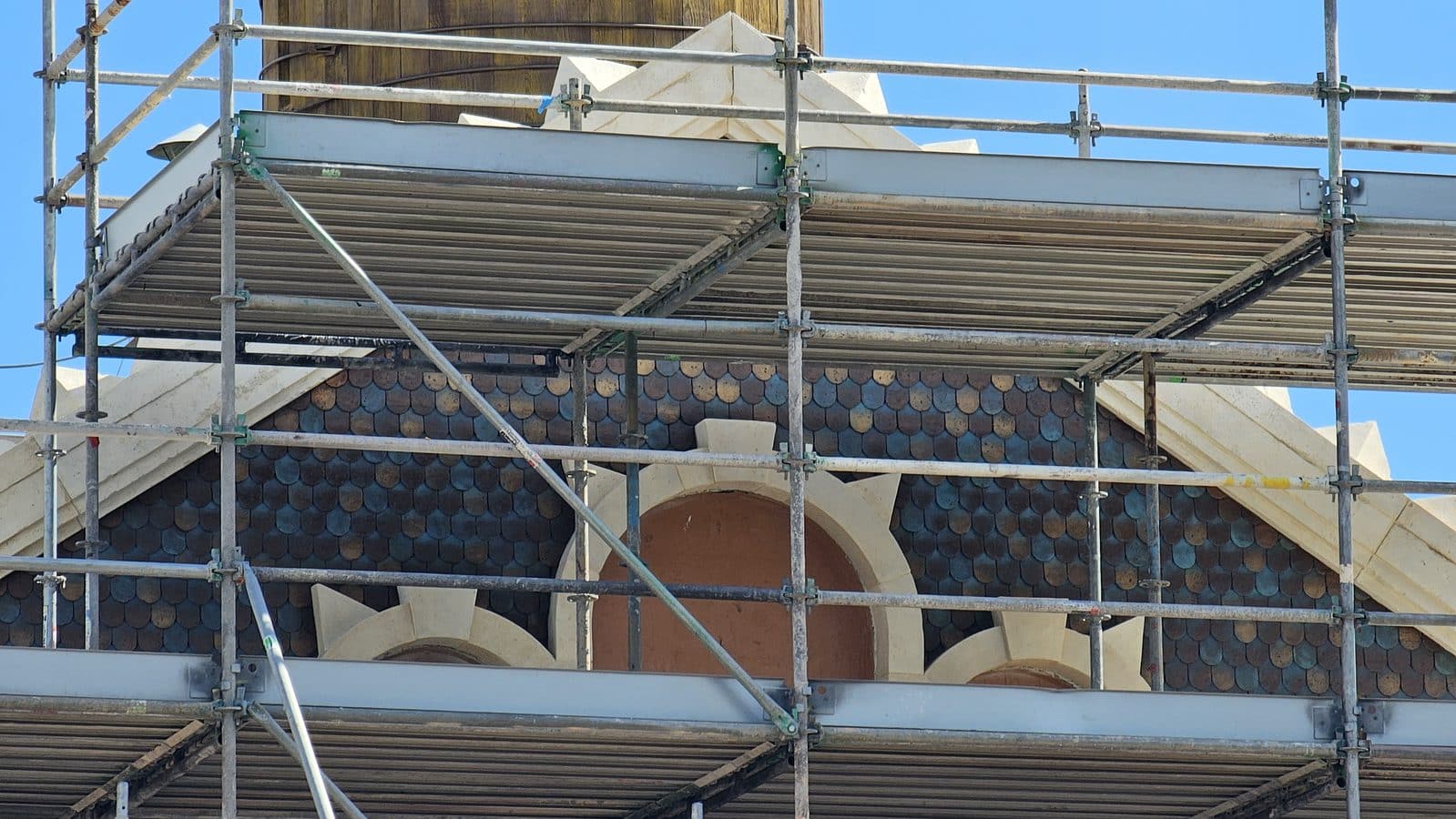 Metal scaffolding surrounds a building facade with a blue tiled roof and arched openings under a clear sky
