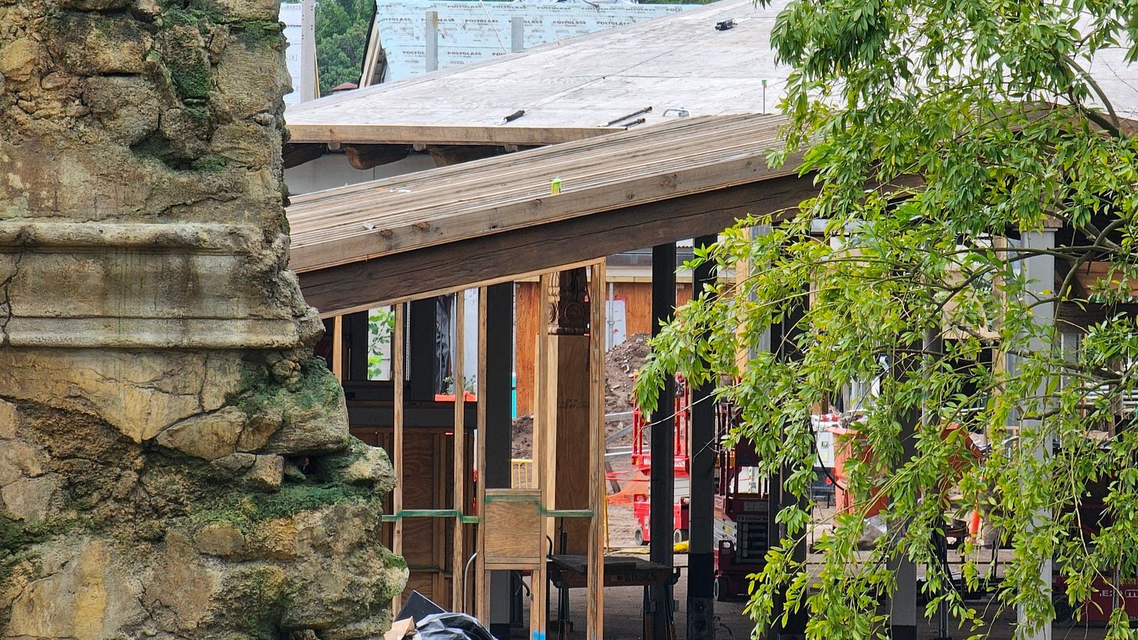 Stone wall on the left side of a construction site with exposed wooden framing and scaffolding under a sloped roof and leafy branches in the foreground on the right