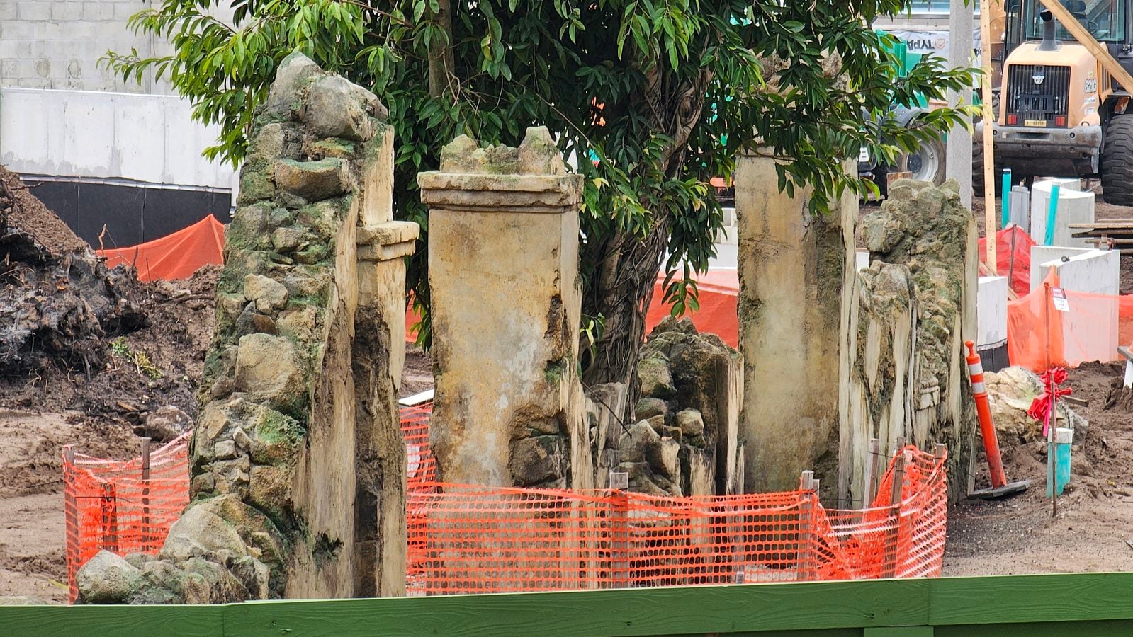 Construction site with mossy stone pillars and orange safety fencing dirt piles in foreground