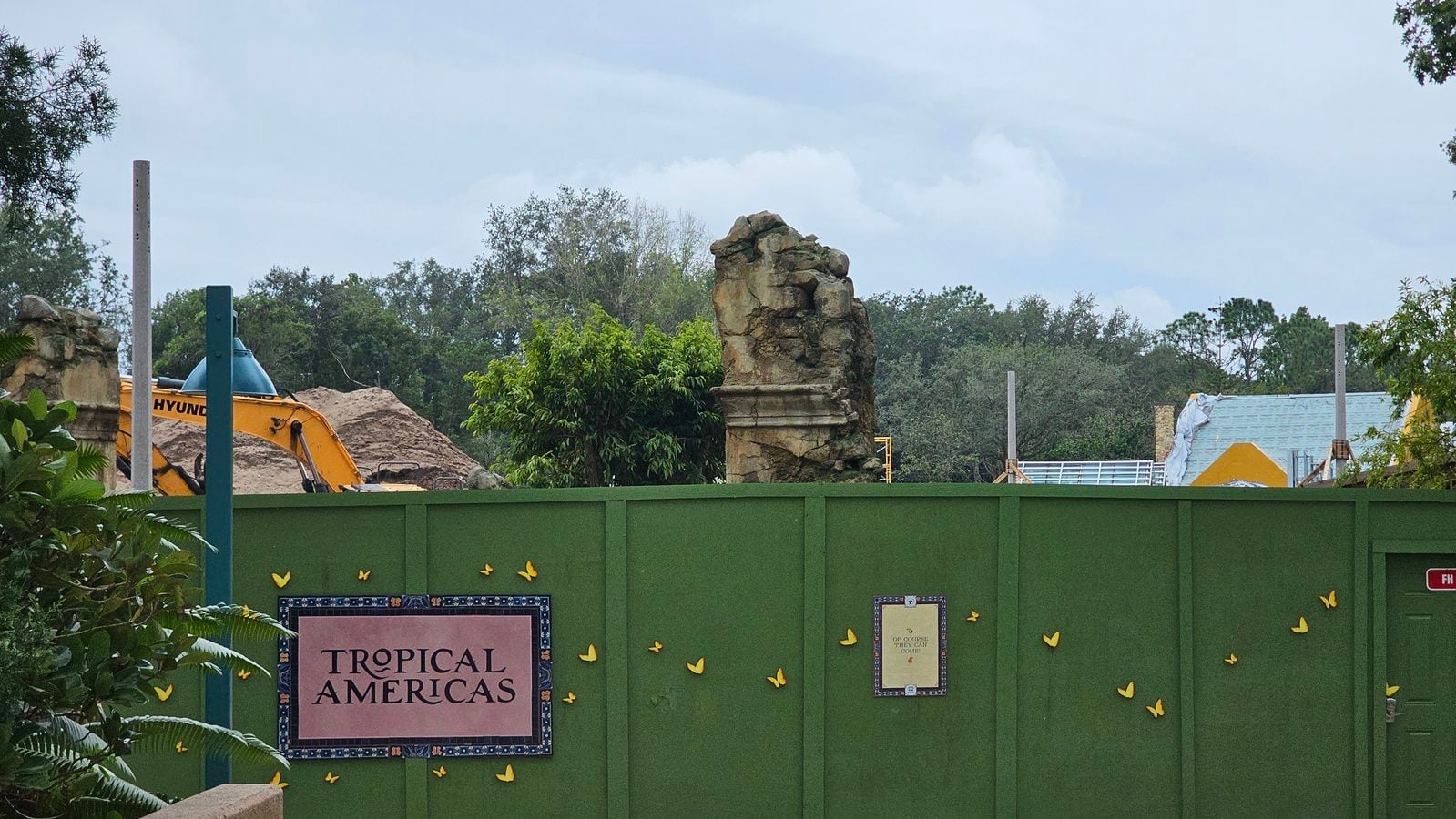 Construction yard behind a tall green fence with a yellow excavator and dirt mounds in the background plus a large rock formation nearby