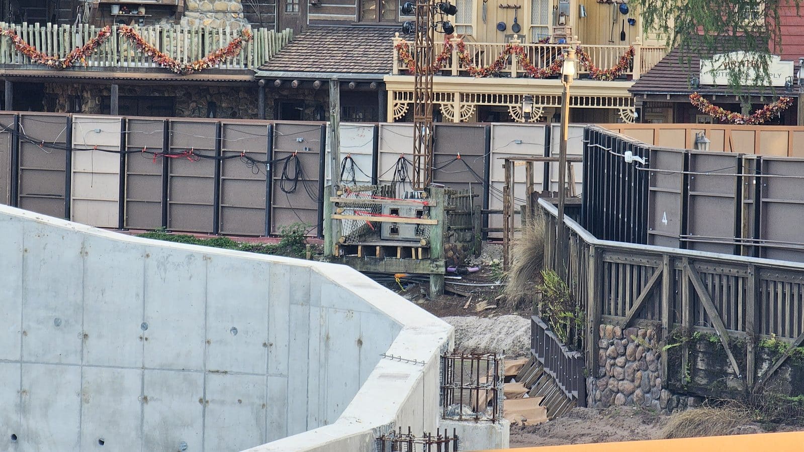 Construction site behind tall gray barriers with scattered cables and a wooden fence and festive garlands hanging along a balcony above A stone and wood building is visible in the background