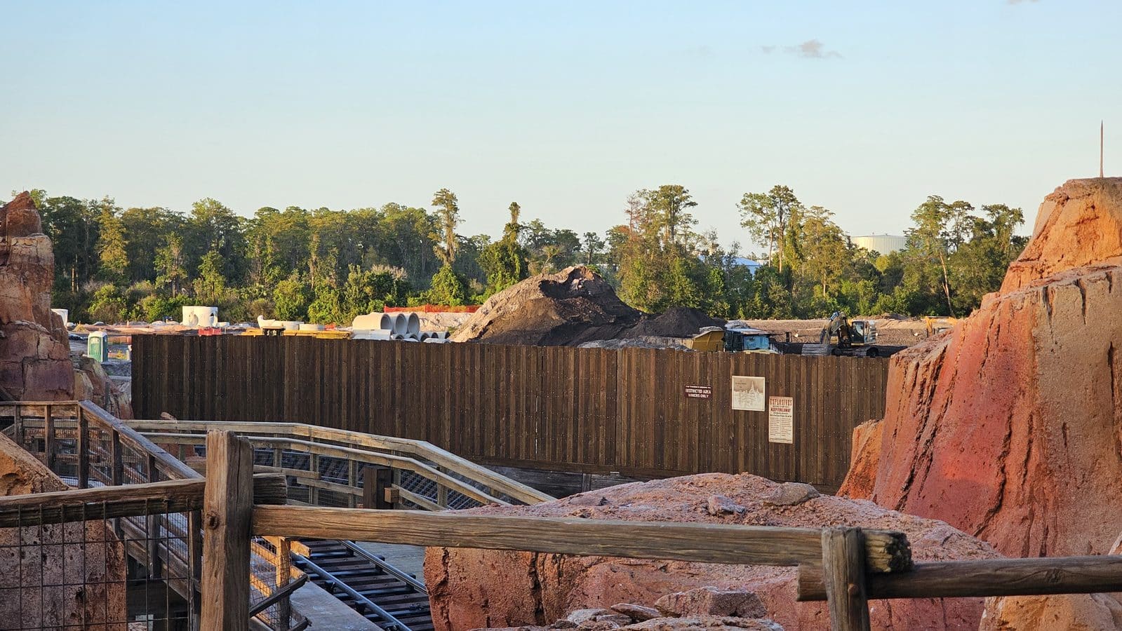 Construction site with dirt piles fencing and a tower of soil against a tree line and blue sky behind