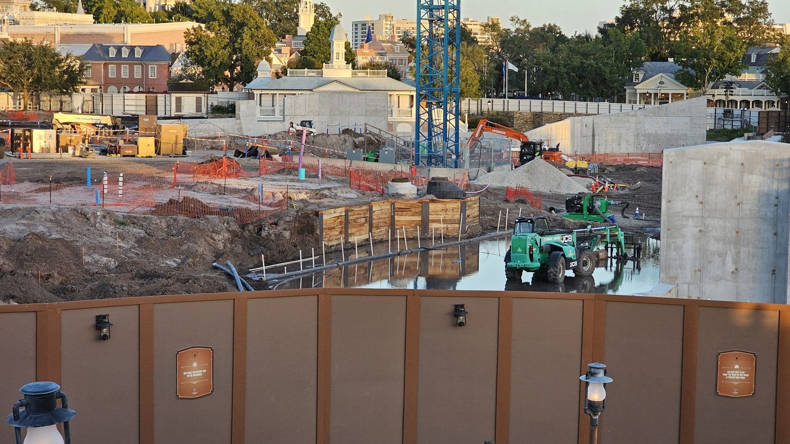 Construction site with heavy machinery excavated earth and a blue crane against a backdrop of buildings and trees