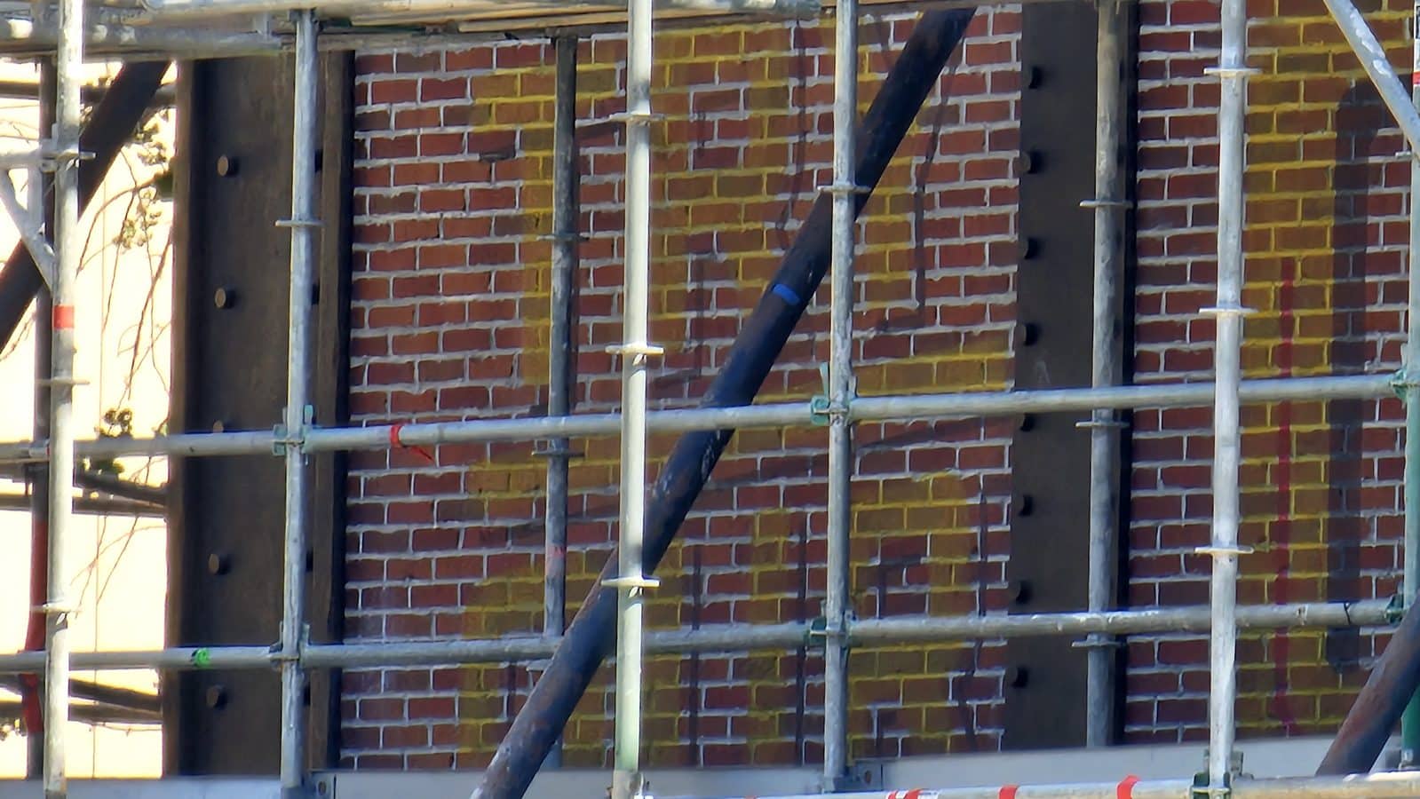 Scaffolding frames in front of a red brick wall at a construction site with metal poles and planks visible