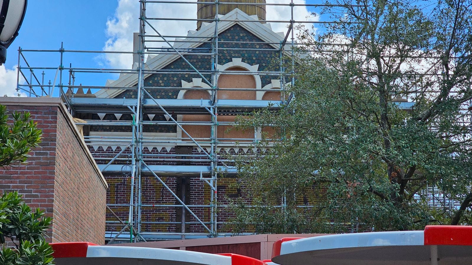 Scaffolding surrounds a decorative brick building with teal slate pattern roof and circular arched openings blue sky above tree to the right