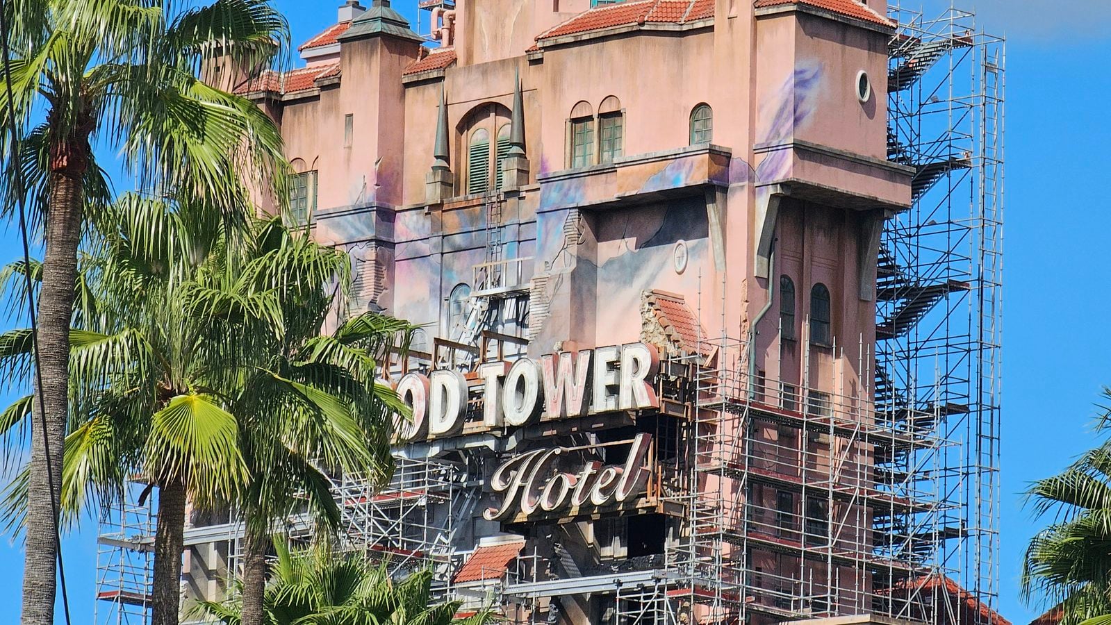 Facade of a vintage pink hotel under scaffolding with a faded neon sign reading Hotel and palm trees against a blue sky