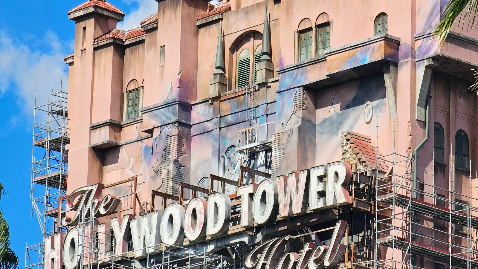 Pink building facade with a large retro style Hollywood sign partially visible amid scaffolding and ladders against a bright blue sky