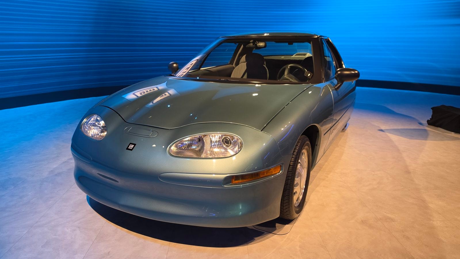 Front three quarter view of a teal blue GM sports car on a showroom floor with a blue backdrop and visible interior