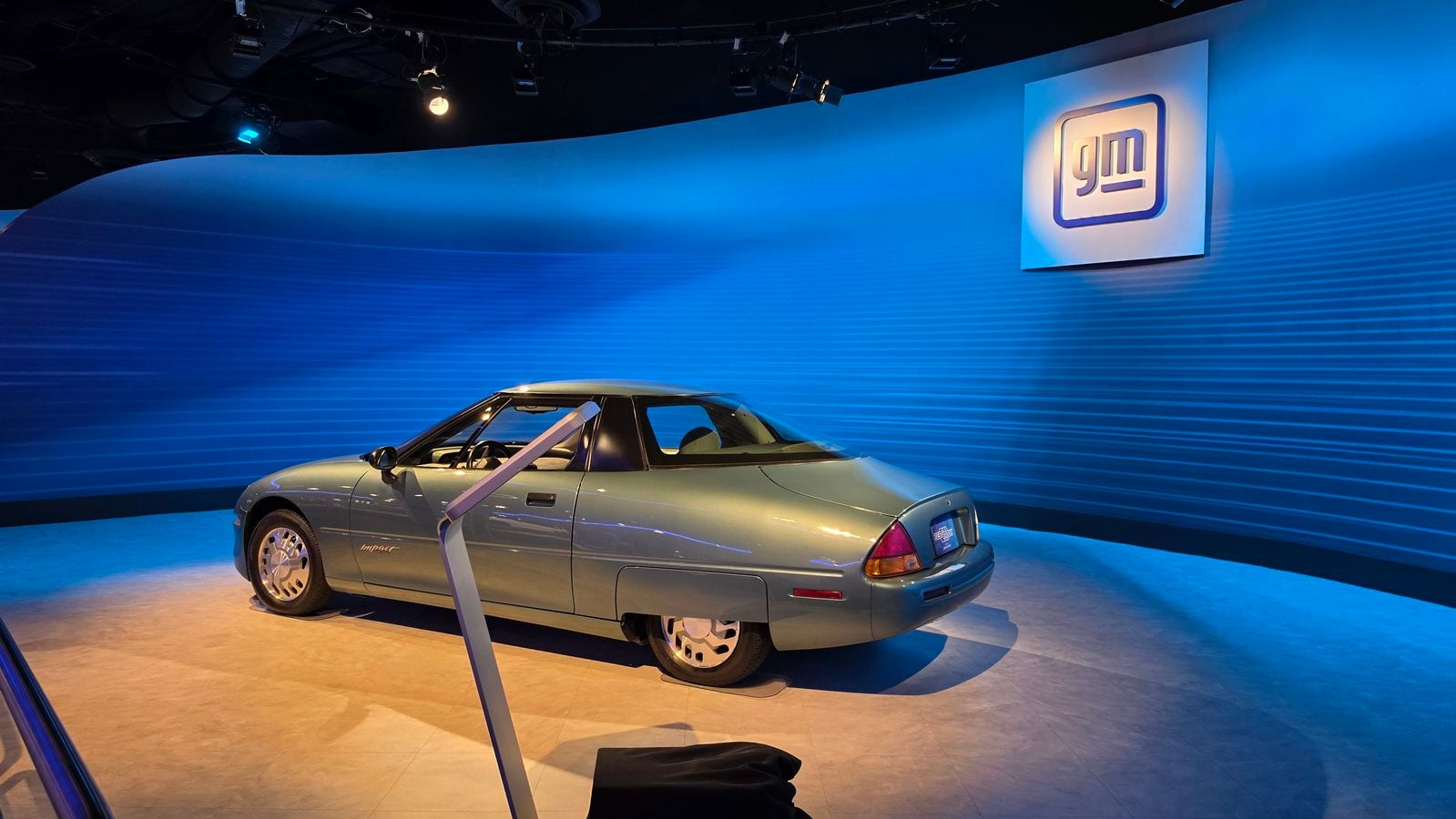 A silver sedan on a showroom floor under blue curved wall lighting with a large square logo in the background