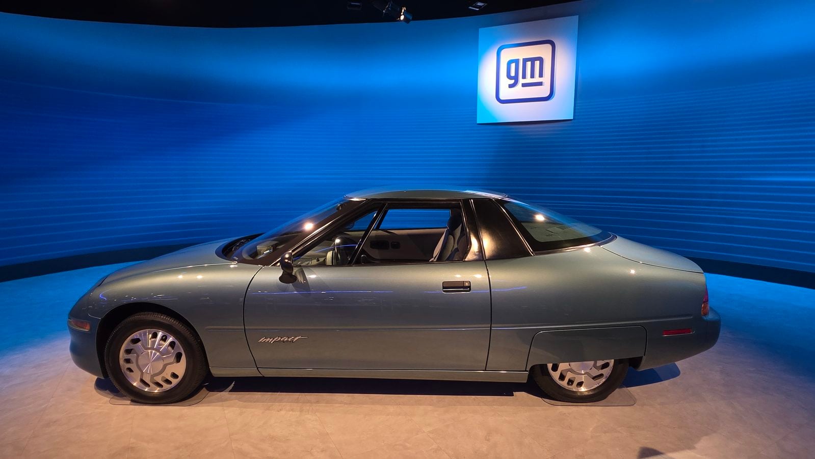 Silver sports car on a showroom floor with a blue curved wall and a lit GM logo above illustrating a display setting