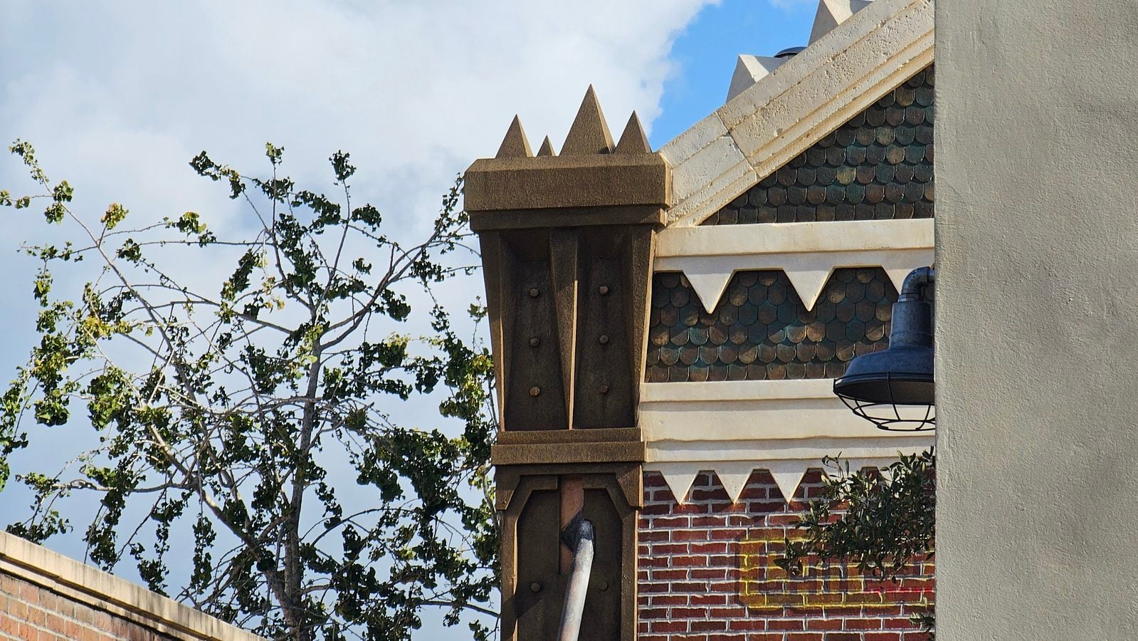 Close up of ornate brick and wooden architectural trim on a building with scalloped roof tiles and a tree against a blue sky in the background