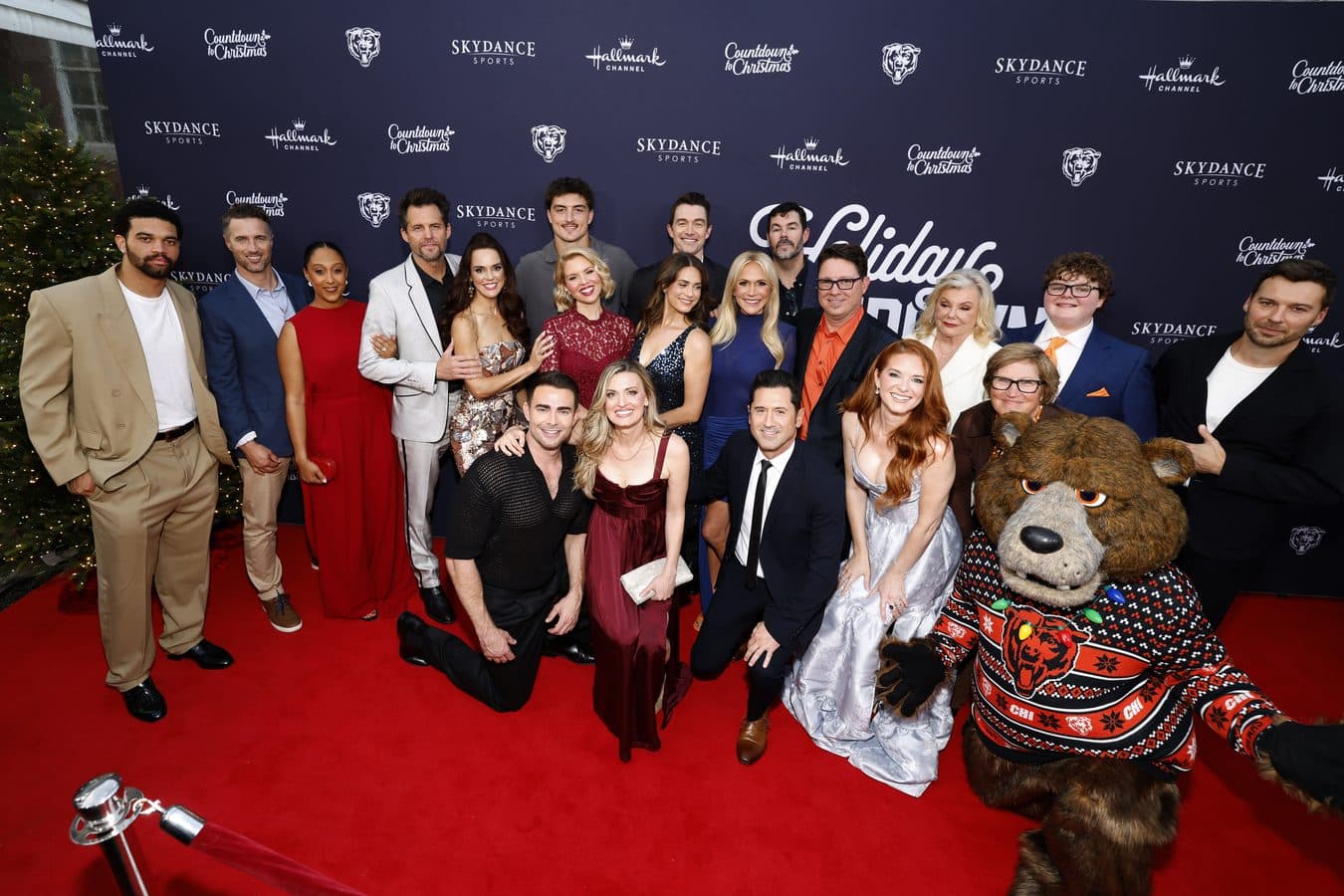 Group photo on a red carpet with guests in formal attire a bear mascot in a festive sweater sits on the right