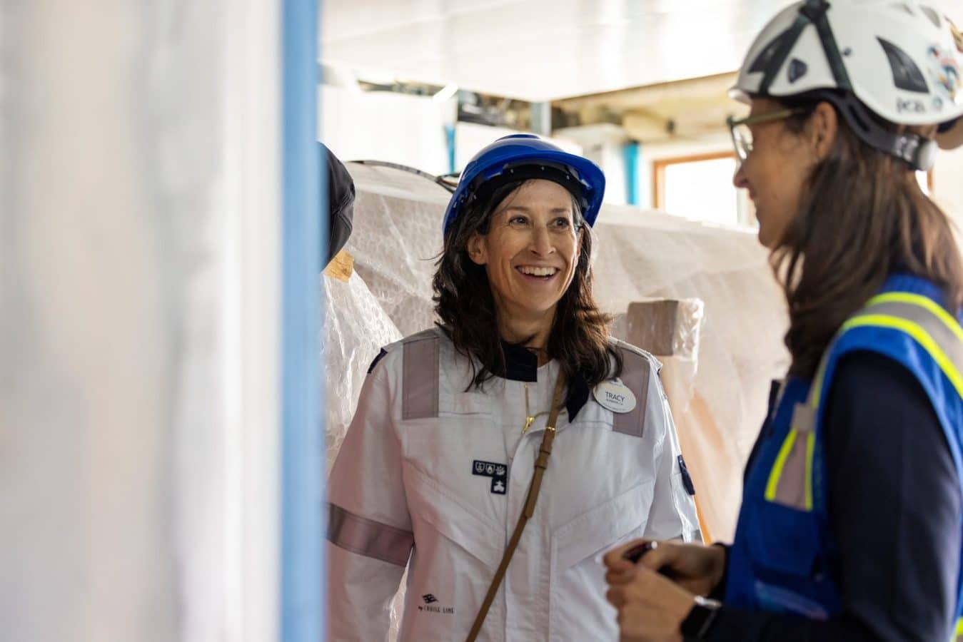 Two women in hard hats and safety vests smile and chat on a construction site