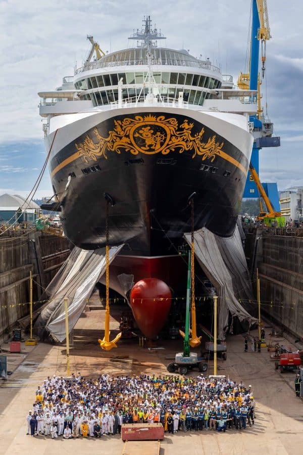 Large cruise ship in a dry dock with ornate gold hull design bow toward camera group of crew and workers posing in front