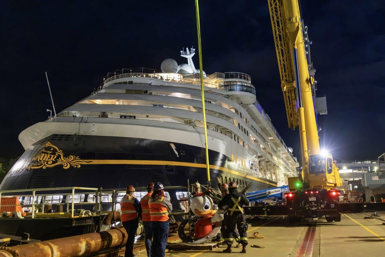 Nighttime scene of the Disney Wonder cruise ship on a dock beside a tall yellow crane with crew in orange vests standing near the hull