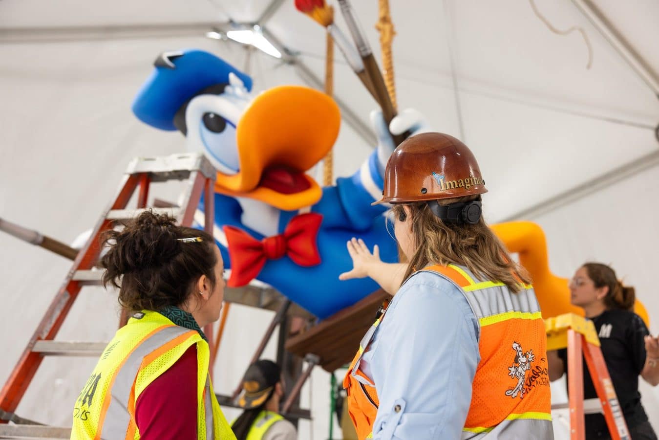 Workers in high visibility vests and hard hats stand around a giant blue duck mascot inside a tent