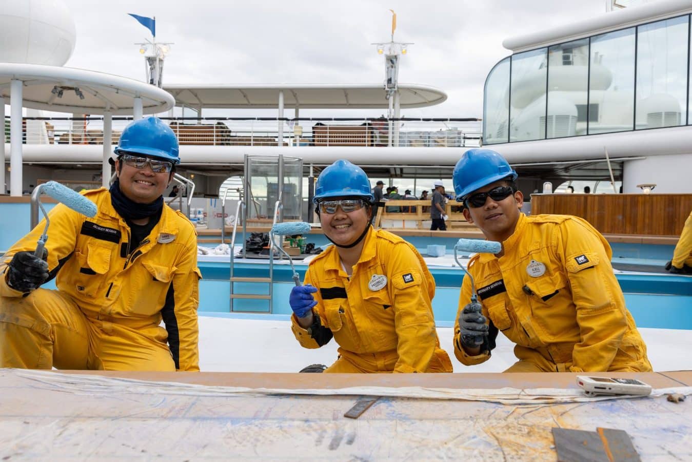 Three workers in yellow coveralls and blue hard hats kneel on a ship deck painting with rollers and smiling at the camera
