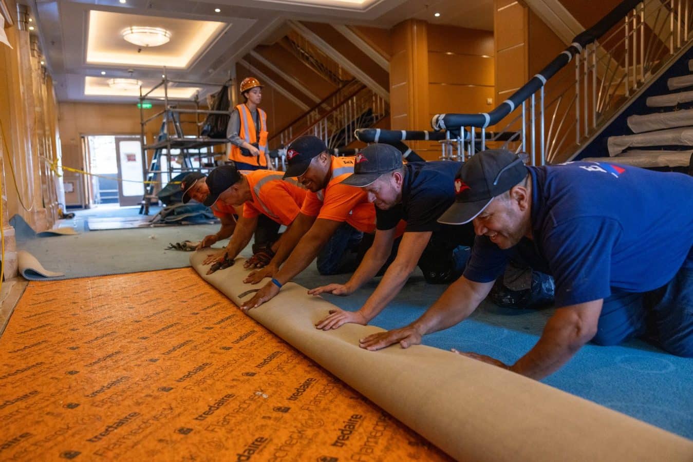 Group of construction workers in orange vests and hard hats kneeling and unrolling floor underlayment in a lobby area