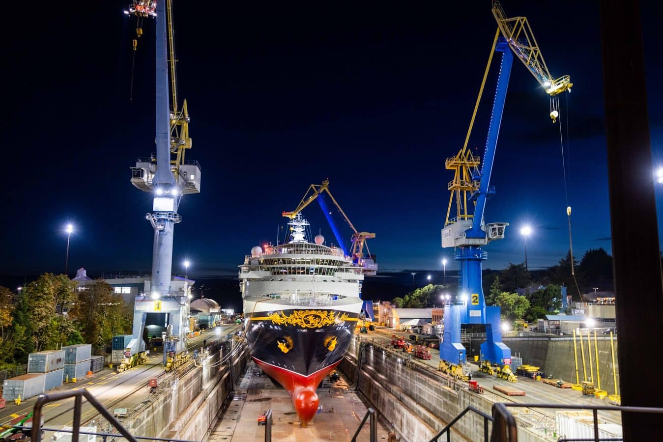Nighttime view of a large passenger ship in a shipyard surrounded by tall cranes and bright work lights