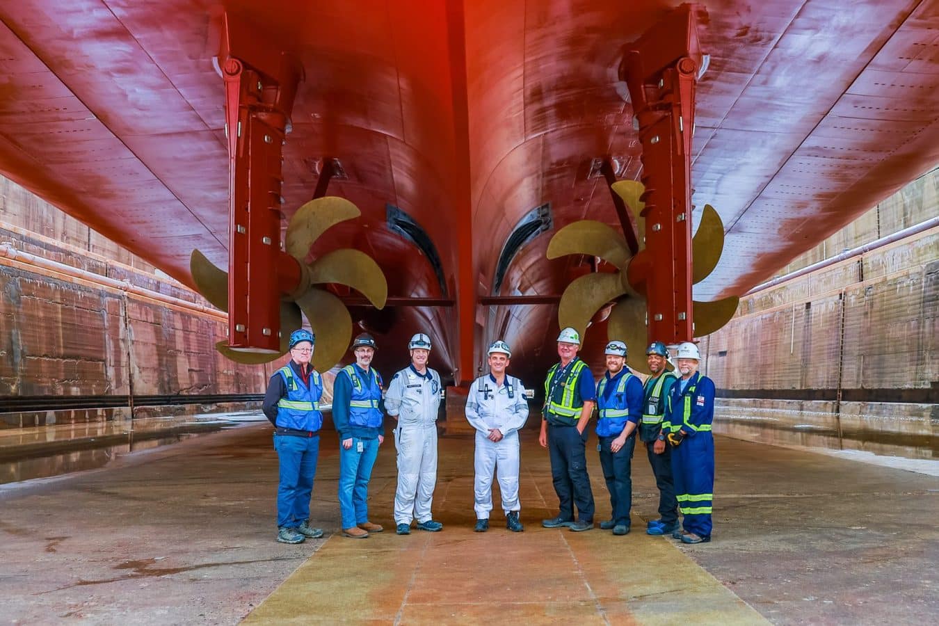 Eight shipyard workers in hard hats and reflective vests stand in a dry dock beside a large red ship propeller