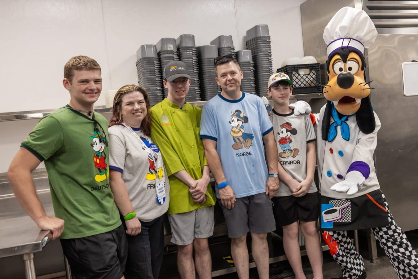 Group of six people in a kitchen posing for a photo with a chef dog mascot in a white coat and hat several wear shirts featuring Mickey Mouse and country names Germany France Norway Canada