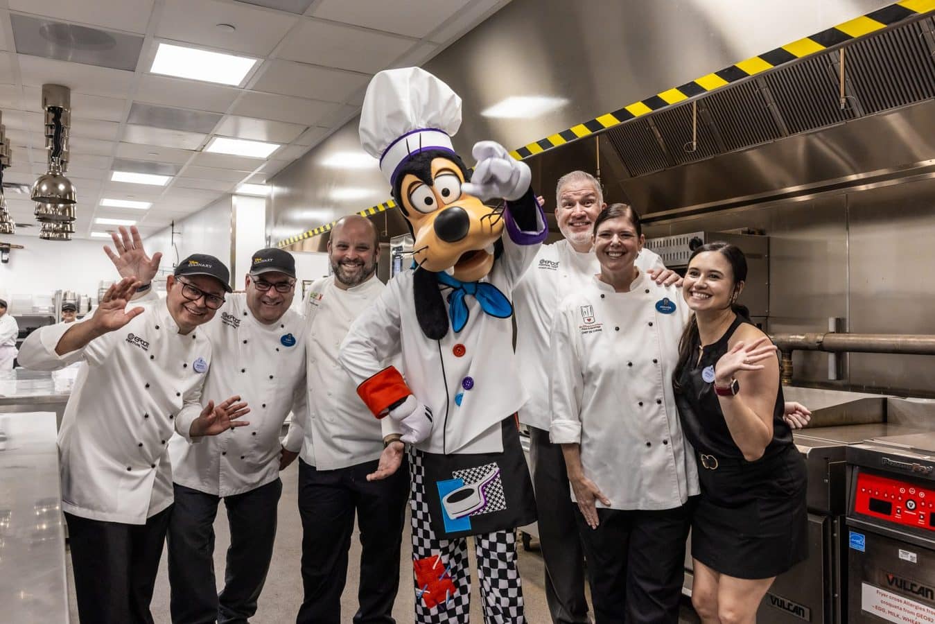 Group of chefs posing with a Goofy mascot in a stainless steel kitchen