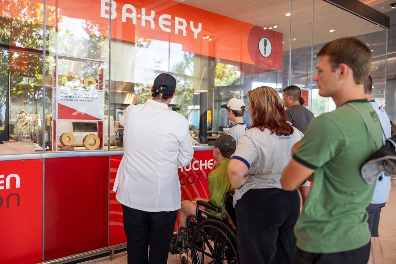 Customers line up at a bakery counter inside a store a staff member in a white coat assists them