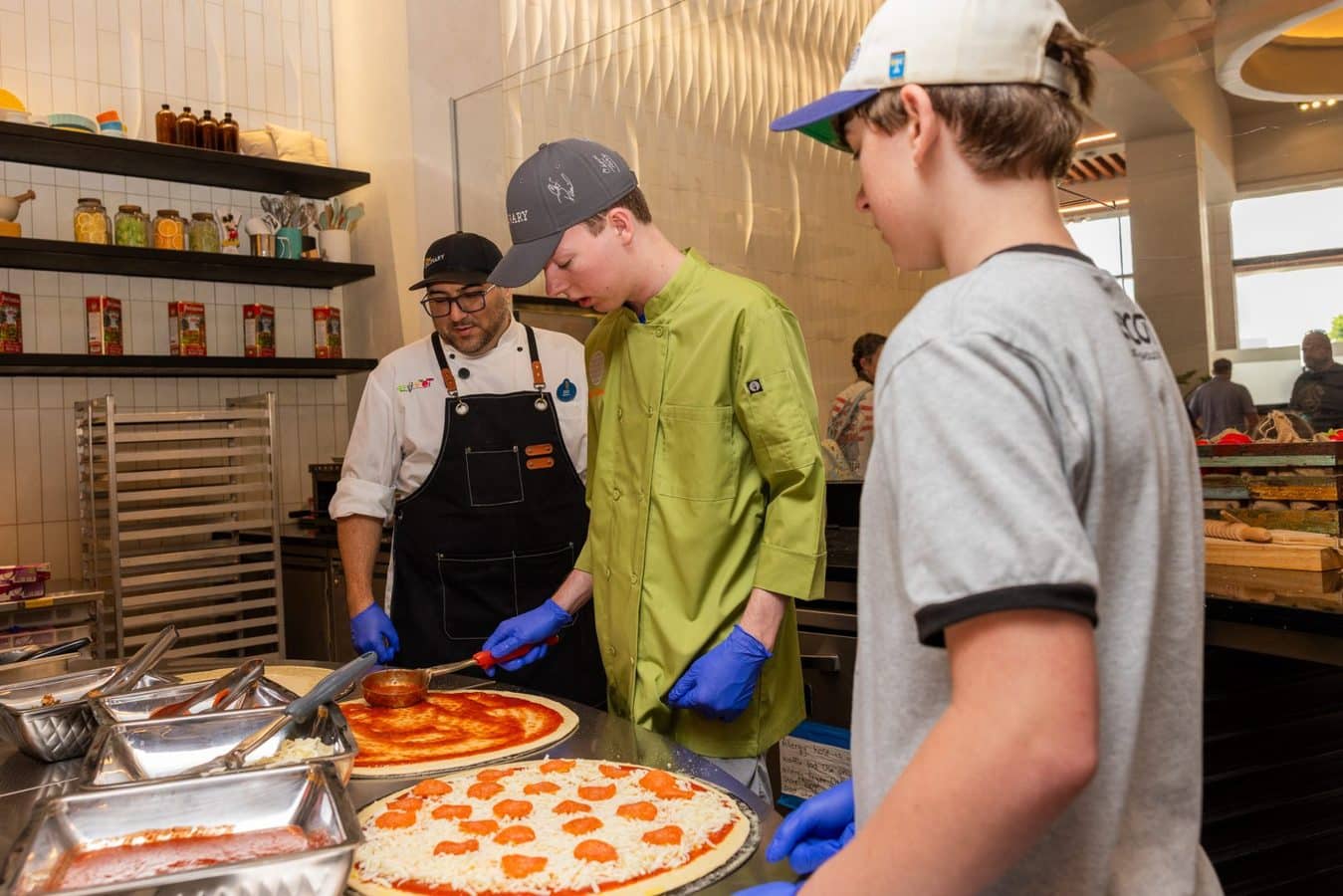 Three people in a modern kitchen prepare pizzas with a pepperoni pizza being spread with sauce on a wooden counter