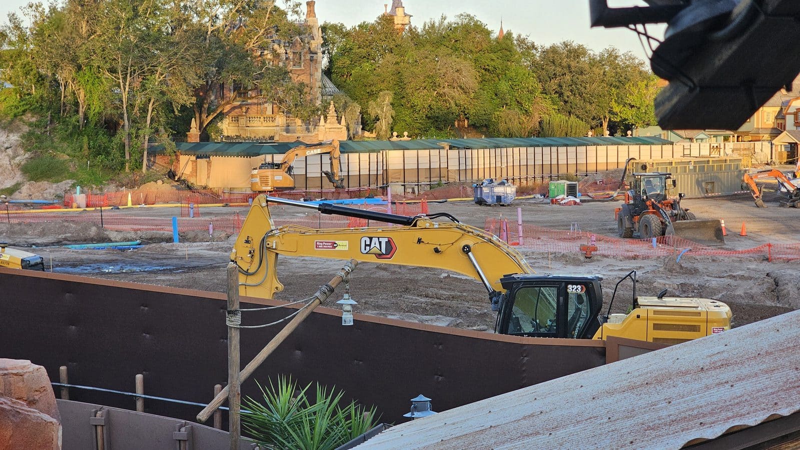 Construction site with a yellow CAT excavator orange safety fencing and a historic building seen through trees in the background