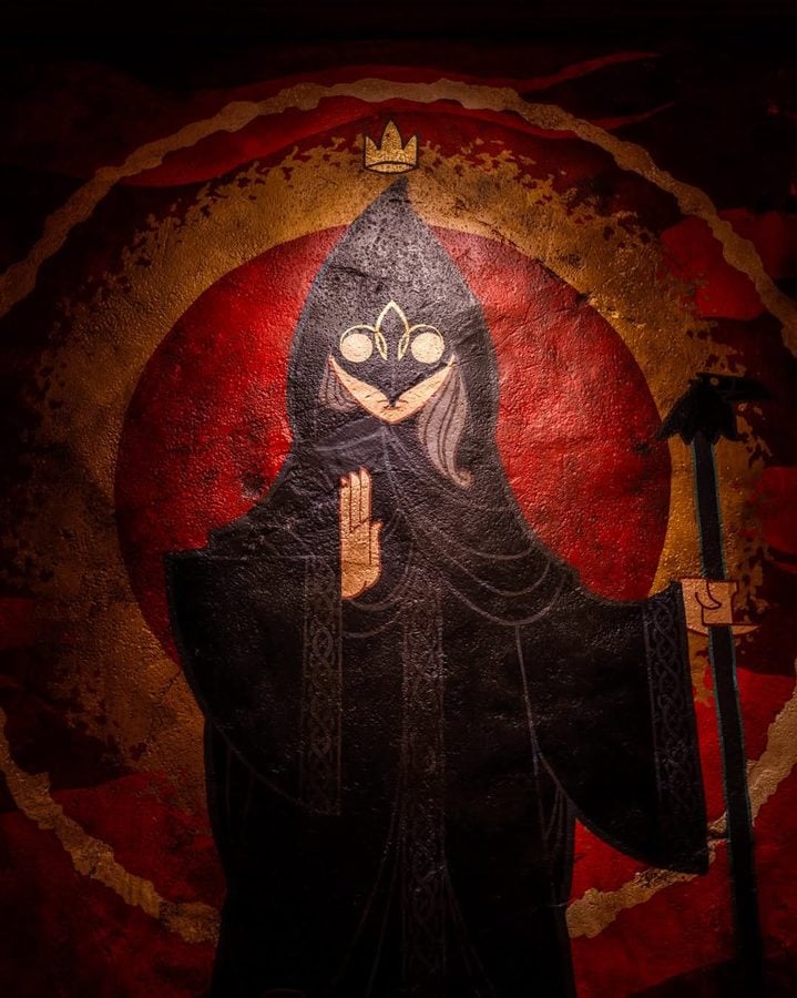 A dark robed hooded figure raises a blessing with one hand wearing a small crown and holding a staff against a red circular halo backdrop